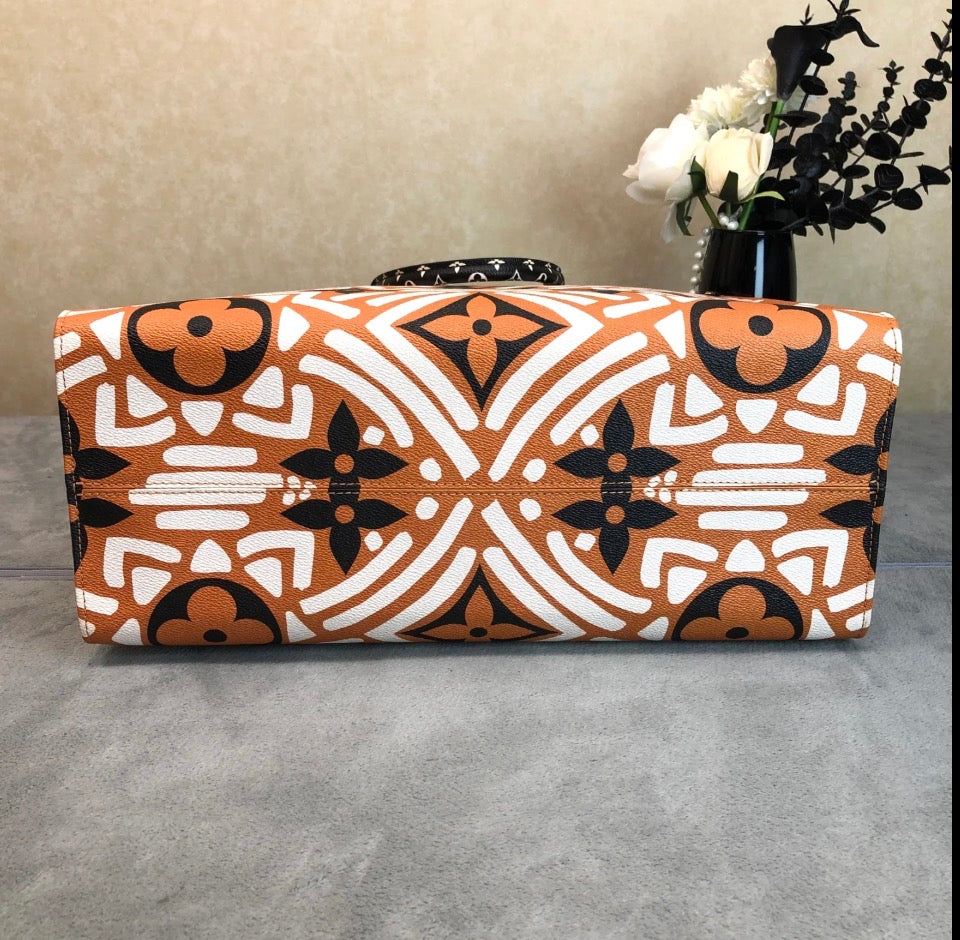 A rectangular handbag with a bold geometric and floral pattern in orange, white, and black, resting on a gray surface with a beige wall and a vase of flowers in the background.