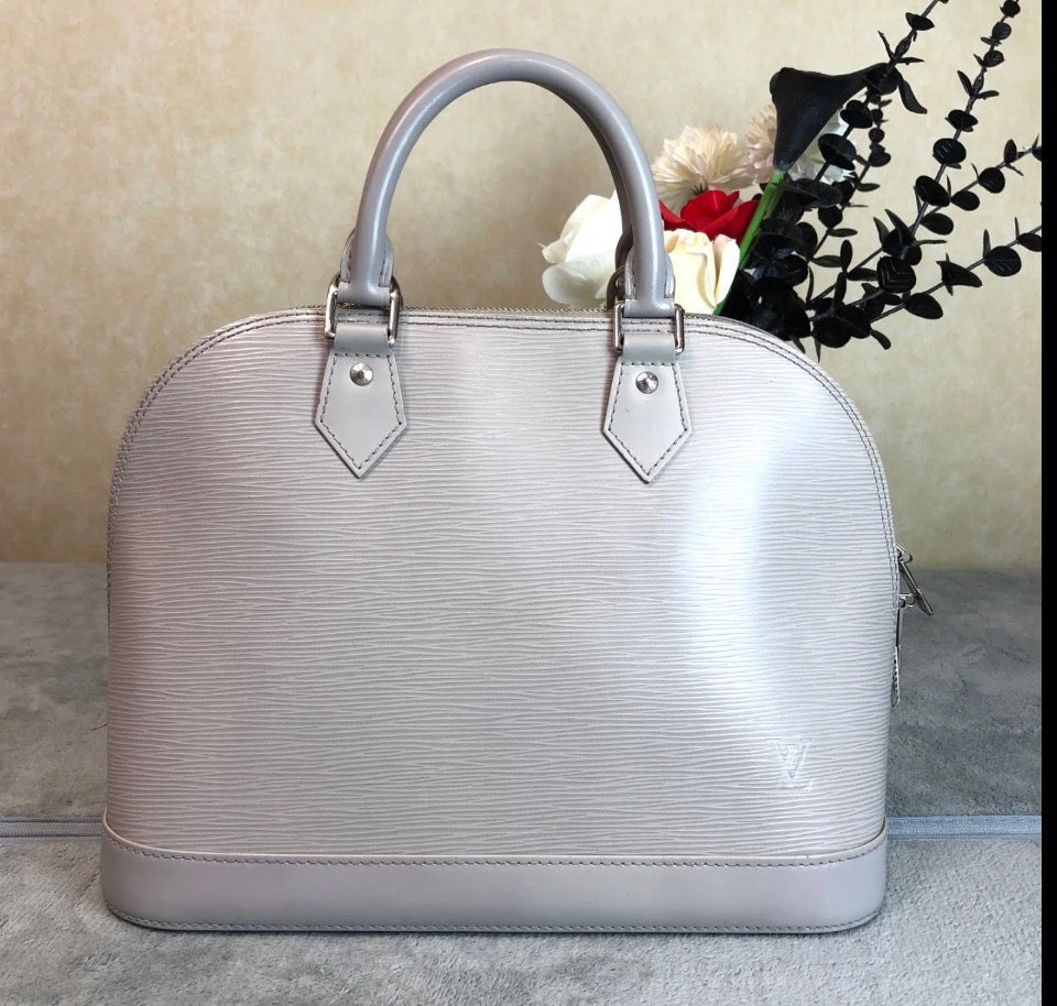 A light silver Louis Vuitton Alma BB handbag with textured Epi leather, featuring dual top handles and a small LV logo on the lower right front, set against a neutral beige background with decorative flowers.