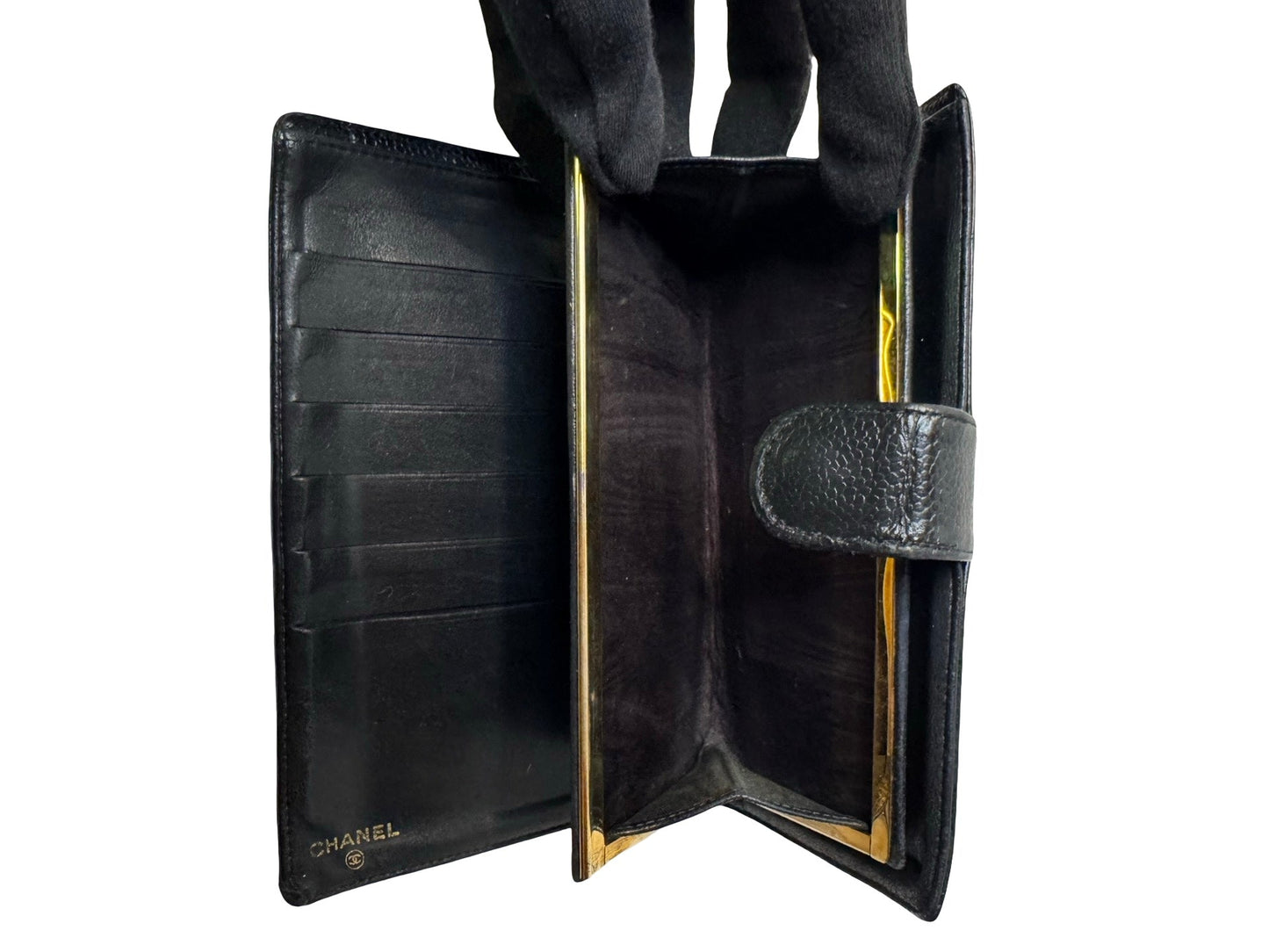 A black leather clutch wallet shown open, revealing its interior compartments and gold-tone metal trim. A gloved hand is visible on the left holding the wallet open.