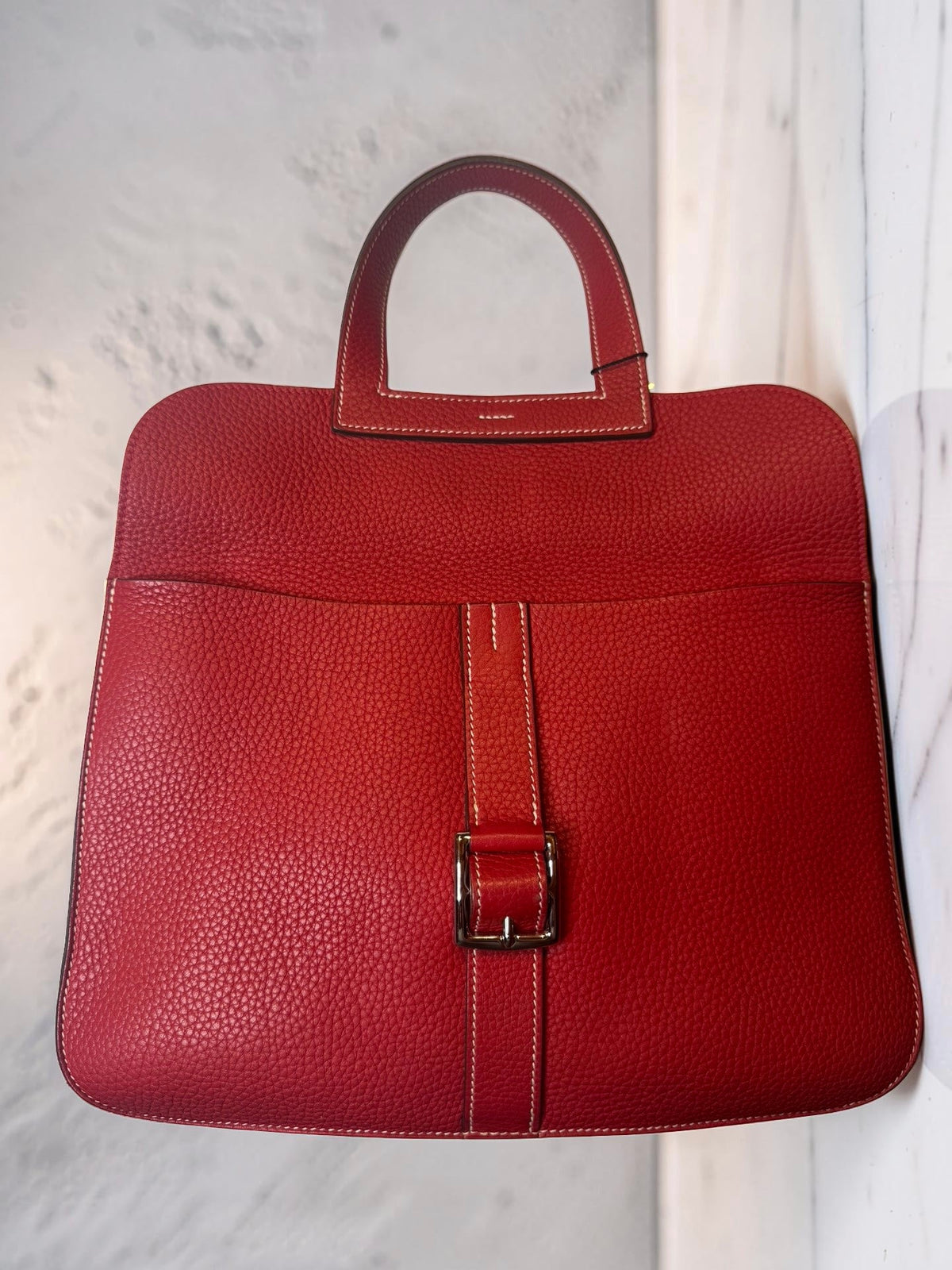 A red pebbled leather tote bag with a top handle and a horizontal strap detail with a silver buckle, set against a light marble background.